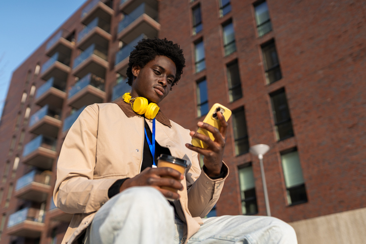Young black man with yellow wireless headphones absorbing content on smartphone while holding coffee cup downtown in the city