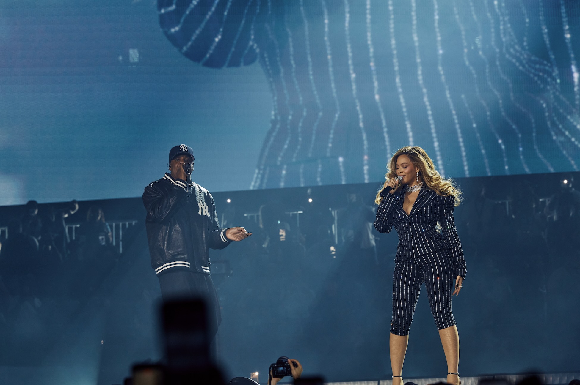Two performers on stage, a man in a Yankees baseball cap and a woman in a black and white striped outfit, singing and dancing under colorful lighting.