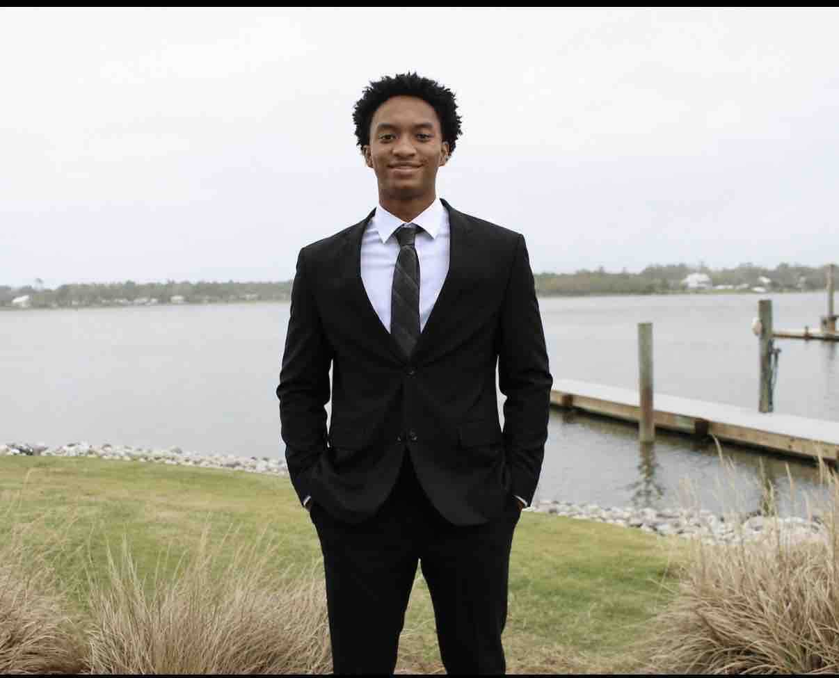 A young man in a black suit and tie standing on a grassy bank overlooking a body of water.