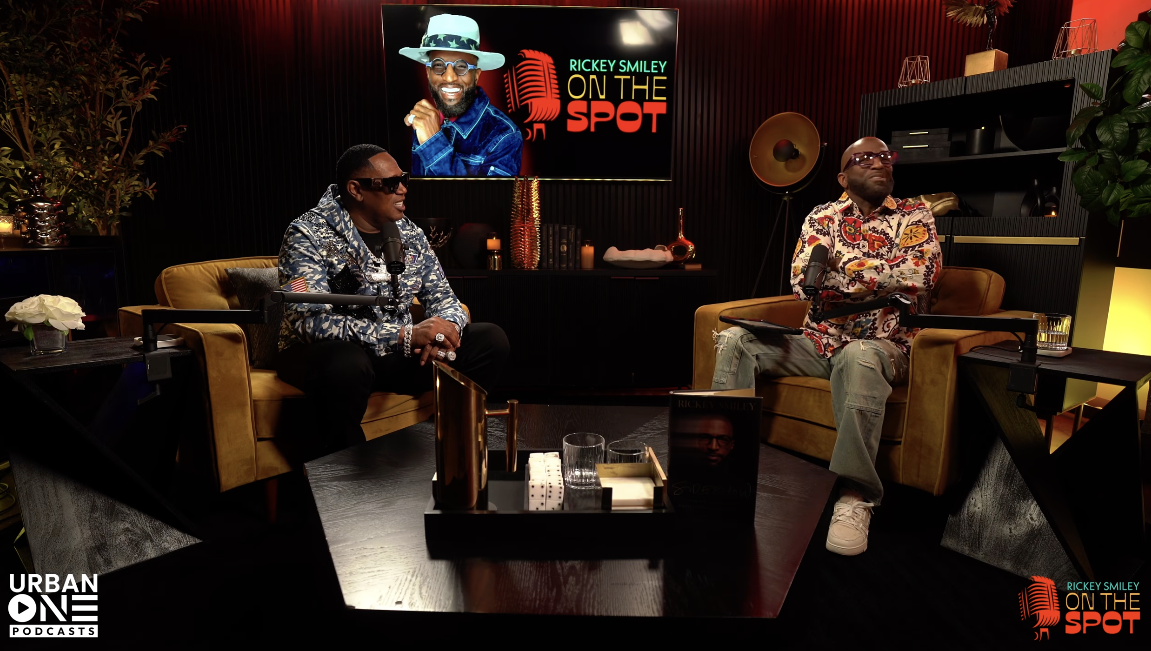 Two men sitting on a couch in a studio setting, with a TV screen displaying
